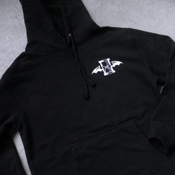 close up, angled image of a black pullover hooded sweatshirt laid flat on a concrete floor. the hoodie has a small white embroidered right chest design of an hourglass with bat wings. 