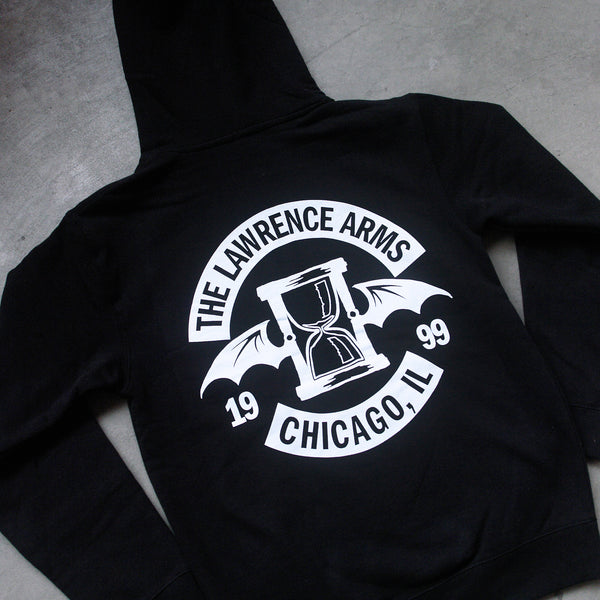 close up, angled image of the back of a black pullover hooded sweatshirt laid flat on a concrete floor. the hoodie  has a full back print in white of the hourglass with bat wings. arched above that says the lawrence arms, and arched up below says chicago, IL