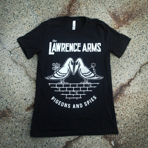  Image of the front of a tshirt against a grey cracked asphalt background. The front of the shirt across the chest in white text reads the lawrence arms. Below that is a graphic of two pigeons face to face with evil smiles on their faces. One is holding flowers behind its back, the other a bomb. They are standing on bricks. Below this in white text reads "pigeons and spies".