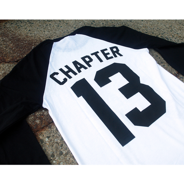 Image of the back of a black and white baseball tee lying against a grey speckled background. The back of the shirt says Chapter 13 in black text. the 13 is printed largely, like a baseball jersey style.