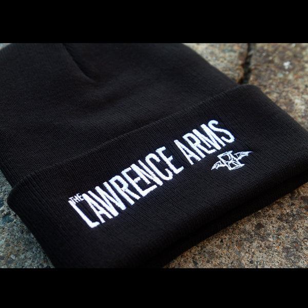 close up Image of a black winter beanie against a grey asphalt background. The beanie folds up at the bottom and on the folded part in white text says the lawrence arms. Under the word arms is the lawrence arms logo of an hourglass with wings.