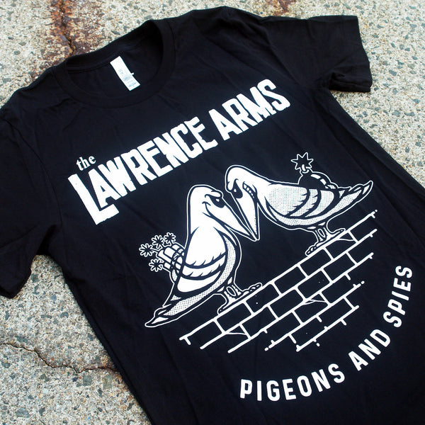 close up  Image of the front of a tshirt against a grey cracked asphalt background. The front of the shirt across the chest in white text reads the lawrence arms. Below that is a graphic of two pigeons face to face with evil smiles on their faces. One is holding flowers behind its back, the other a bomb. They are standing on bricks. Below this in white text reads "pigeons and spies".