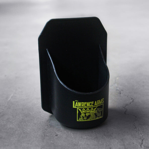 image of a black. plastic shower beer holder on a concrete floor. the back is flat and has adhesive that will stick on a shower wall. the front has a round spot for a beverage can. on the front is a small print in yellow of the suicide king with a sword through his head and the words the lawrence arms at the top