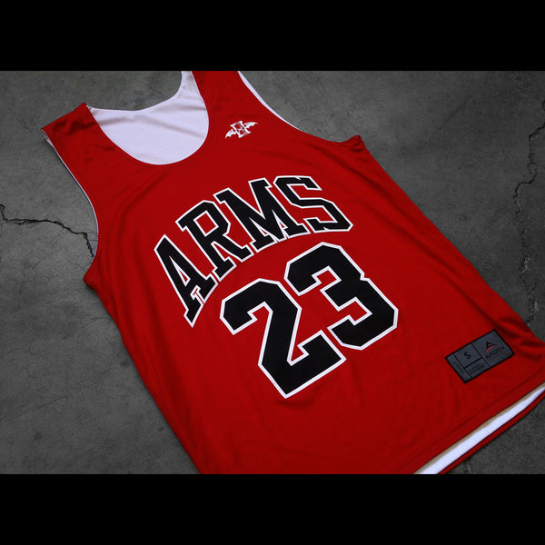 Close up image of a red sleeveless jersey lying against a grey background. The front of it says ARMS across the chest in black with a white outline. Below that is the number 23. A white hourglass with wings the lawrence arms logo is in the upper left chest area. The bottom of the jersey has the brand and size tag on it.