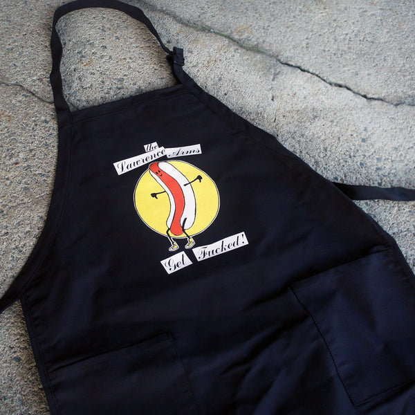 Image of the front of a black apron lying against a grey cracked asphalt background. background. It says "The lawrence arms" in black cursive on top of a white rectangle. Below that is a circle that is filled in with yellow color. Inside of the circle is a graphic of a hot dog with arms and legs and a smiley face on it. Below that it says Get fucked! In black text on top of white rectangles.